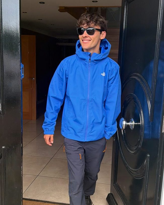 Leo Miller in a blue rain jacket and sunglasses, stepping out of a doorway